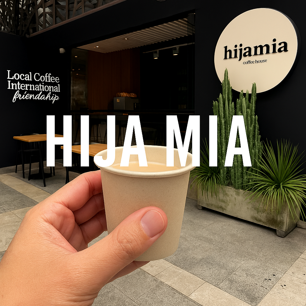 Close-up of a hand holding a paper coffee cup before a modern black café façade featuring a round logo sign, outdoor tables with chairs, and tall cacti.
