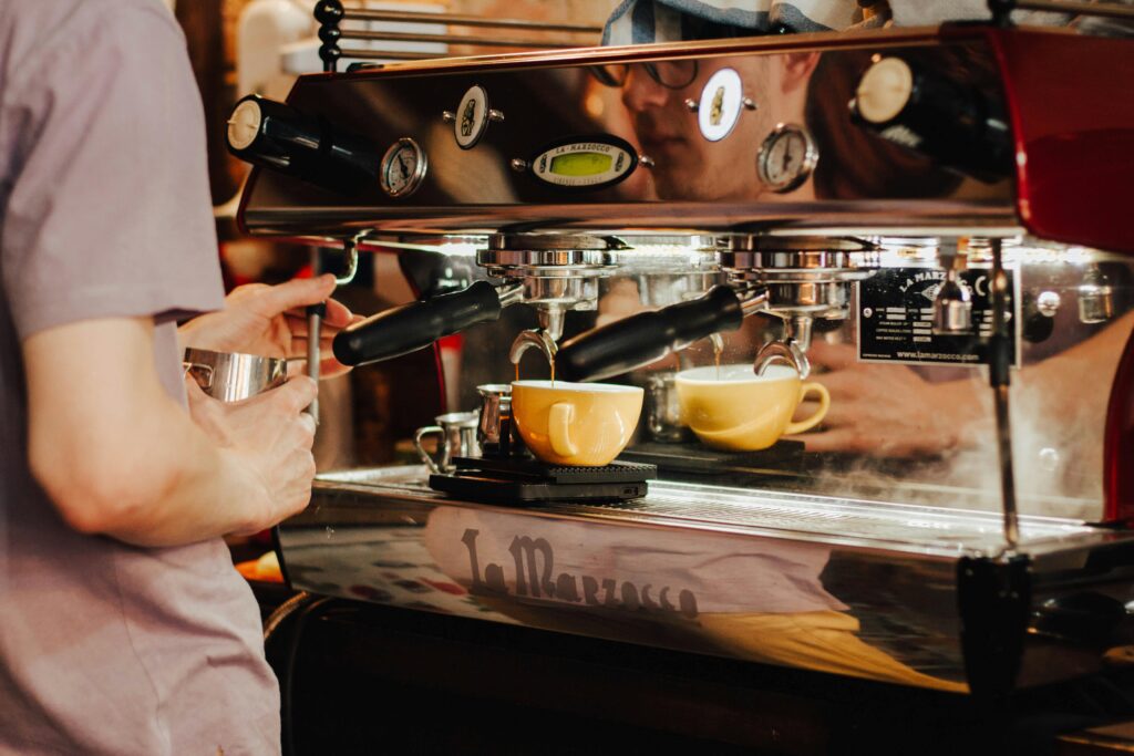 Close-up of a barista using an espresso machine to pull shots into two yellow cups while holding a milk-frothing pitcher.